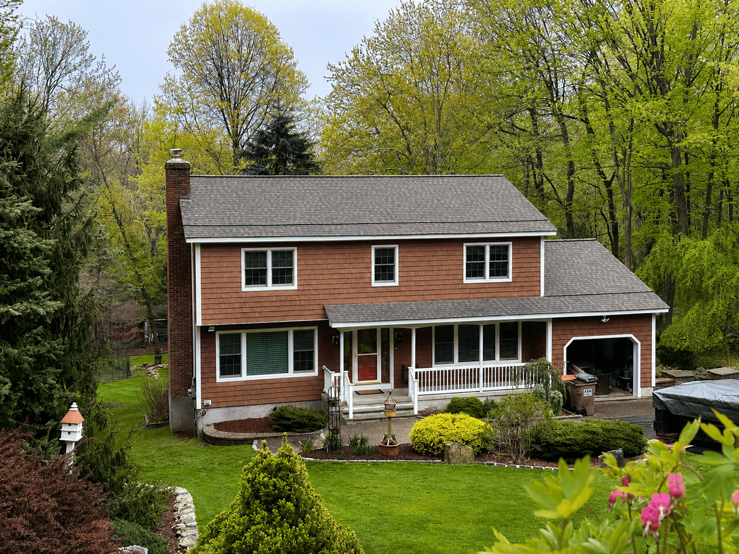 Two-story home clad in cedar-toned shingle siding, with a brick chimney, covered porch and mature landscaping.