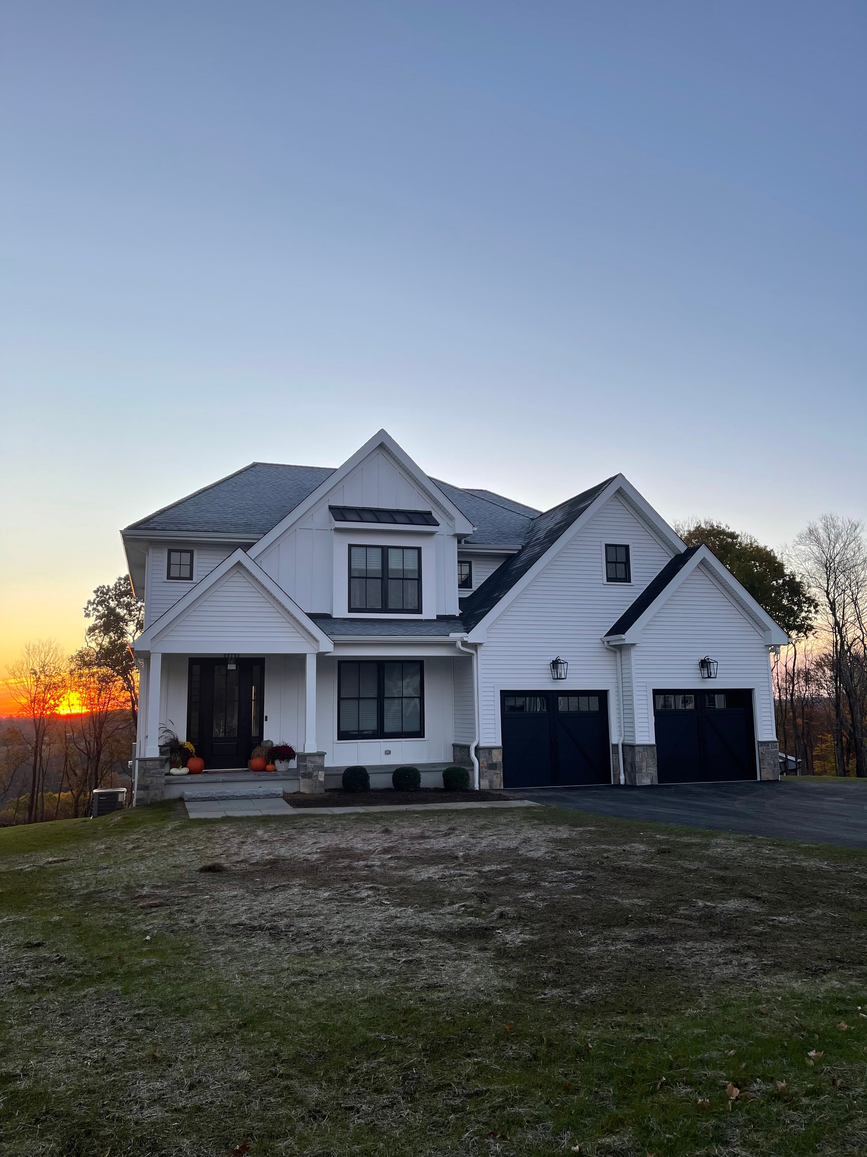 White farmhouse-style home photographed at sunset, with black windows, a covered porch and a two-bay garage with a stone-veneer base.